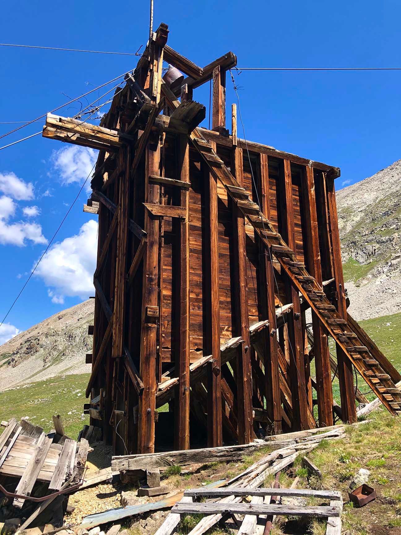 Rusty wooden mining headframe on a rocky hillside under a bright blue sky with clouds. Mayflower Gulch, Colorado