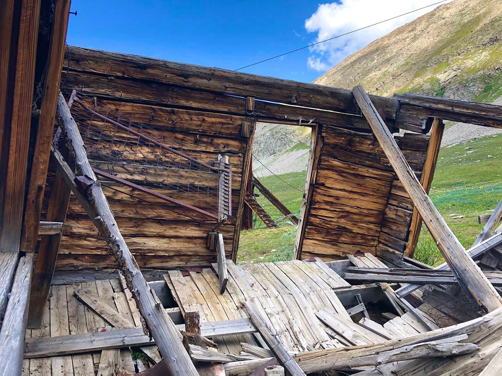 Collapsed wooden shelter with broken roof and walls in a grassy mountain landscape under blue sky, The ruins of an old cabin, Mayflower Gulch, Colorado