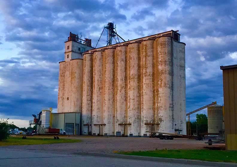 Large grain elevator with tall tan silos under a cloudy blue sky, beside a road and small industrial buildings