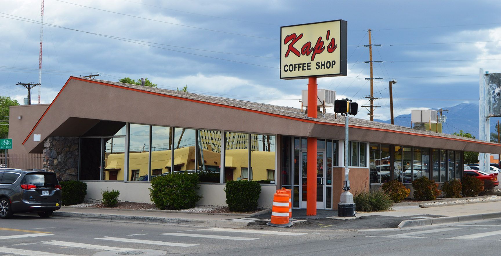 Small roadside restaurant with a red roof, parking lot, and sign by the street under cloudy sky.