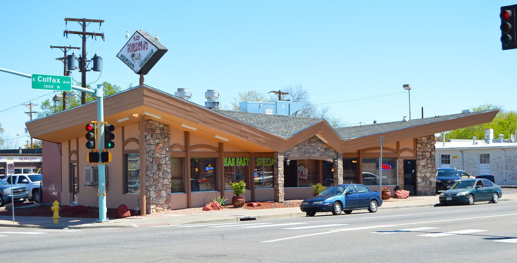 Street-view of a low brick commercial building with a peaked wooden roof, parked cars, and a traffic light at the corner