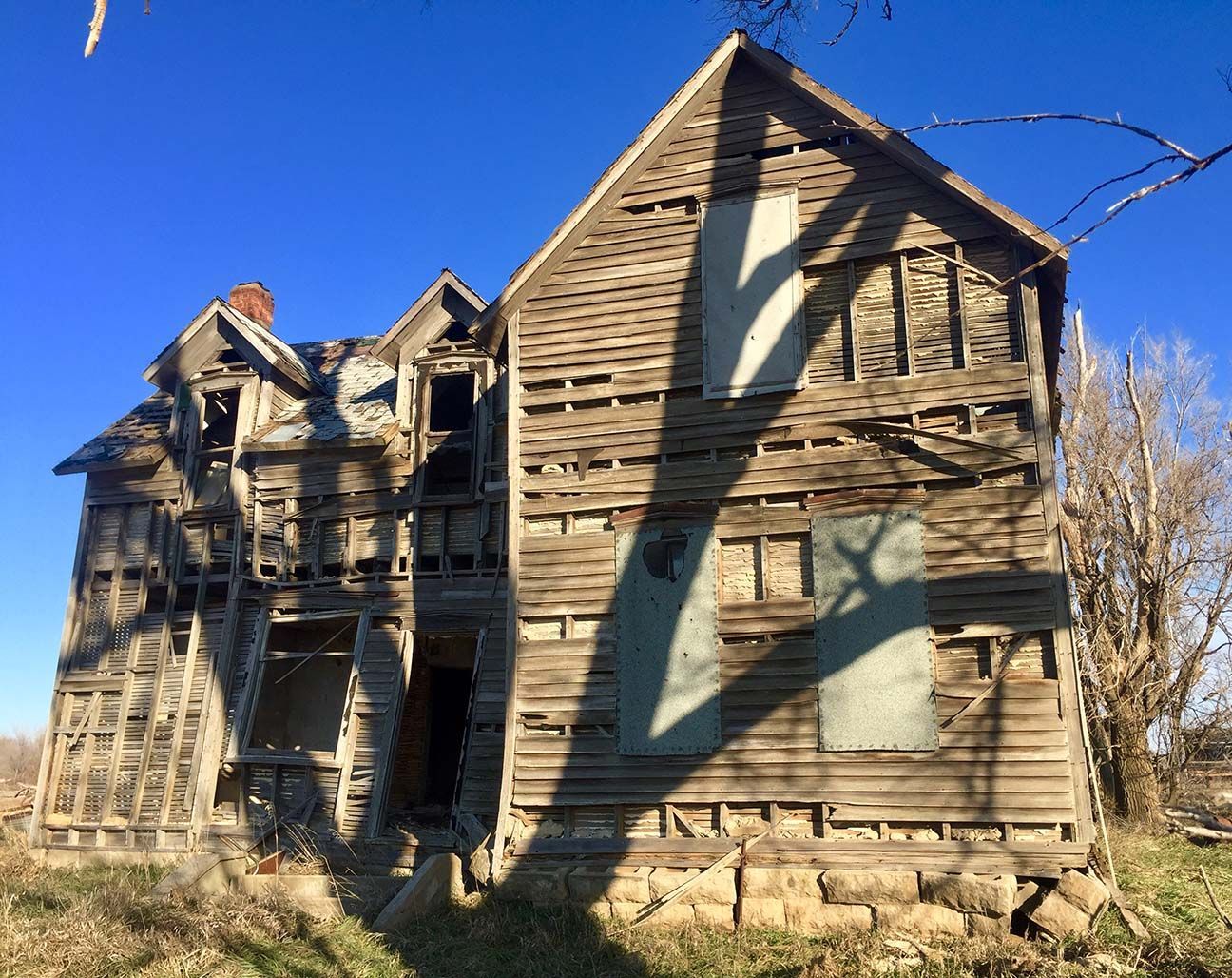 Dilapidated wooden house with peeling siding under a bright blue sky, overgrown yard in front