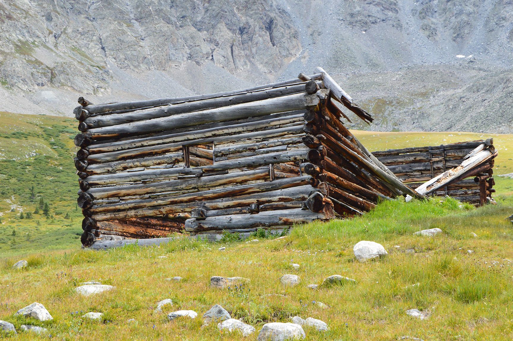 Rustic wooden shelter on a grassy hillside beneath a rocky mountain slope. The ruins of an old cabin, Mayflower Gulch, Colorado