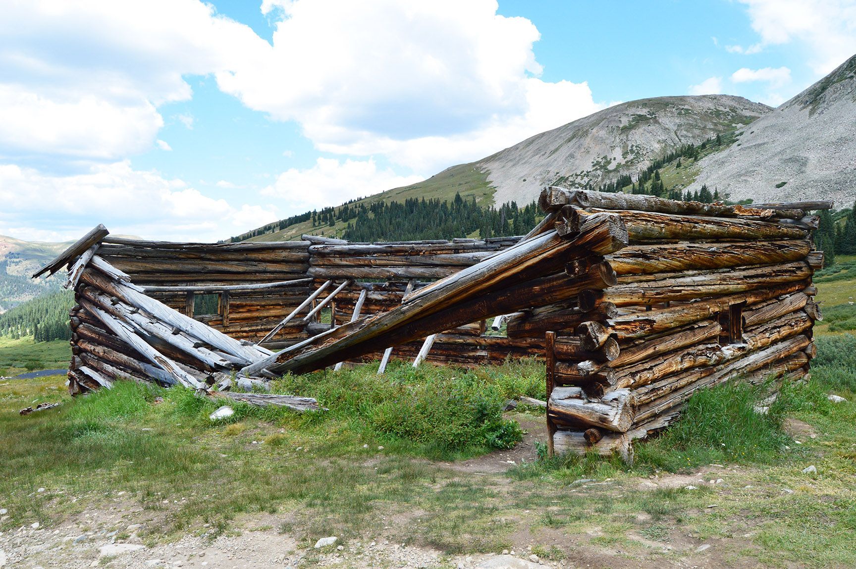 Collapsed log cabin in a grassy field with mountains and clouds in the background. The ruins of an old cabin, Mayflower Gulch, Colorado