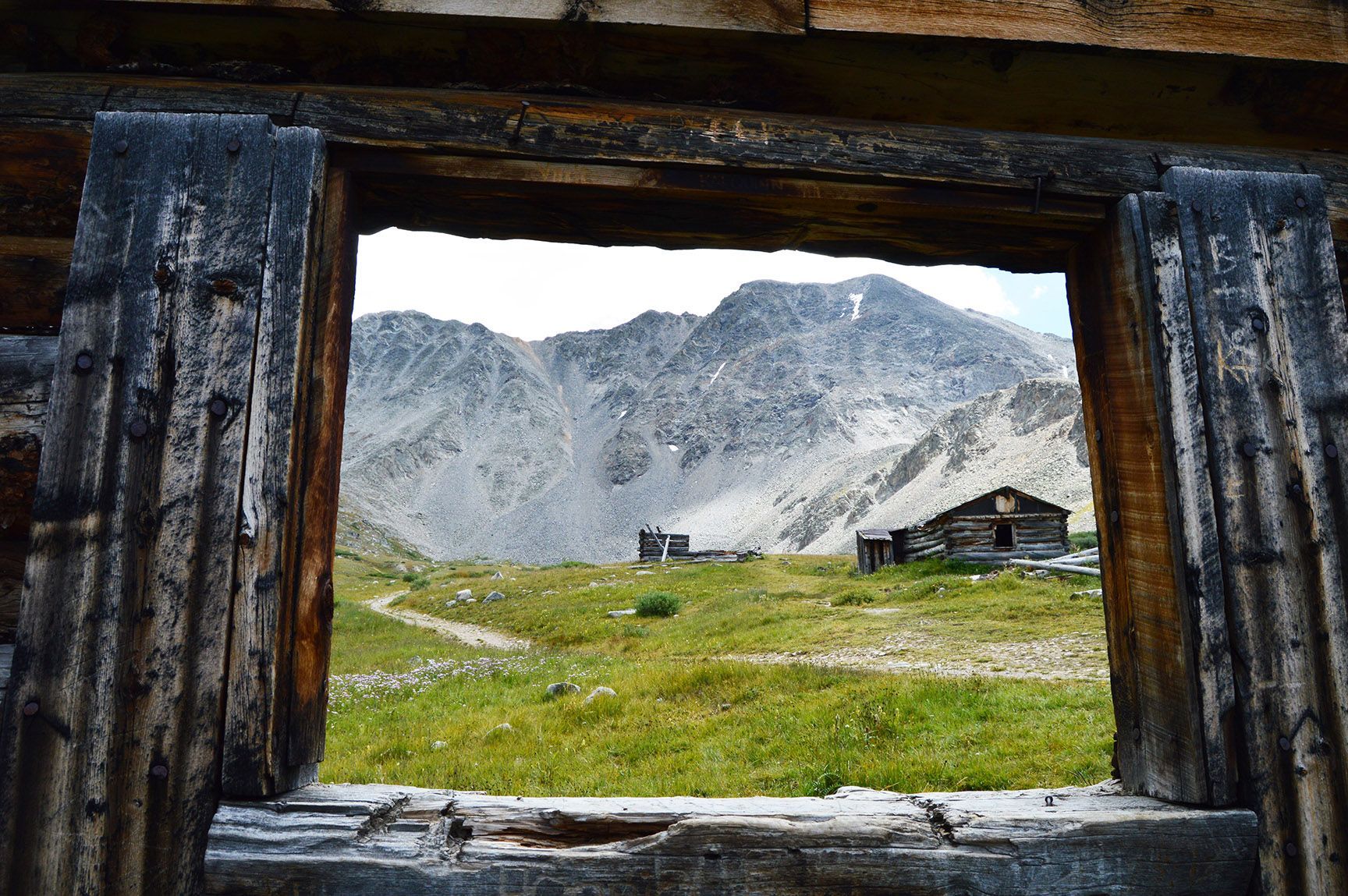 Mountain meadow seen through a rustic wooden window frame, with rocky peaks and small buildings in the distance, The ruins of an old cabin, Mayflower Gulch, Colorado