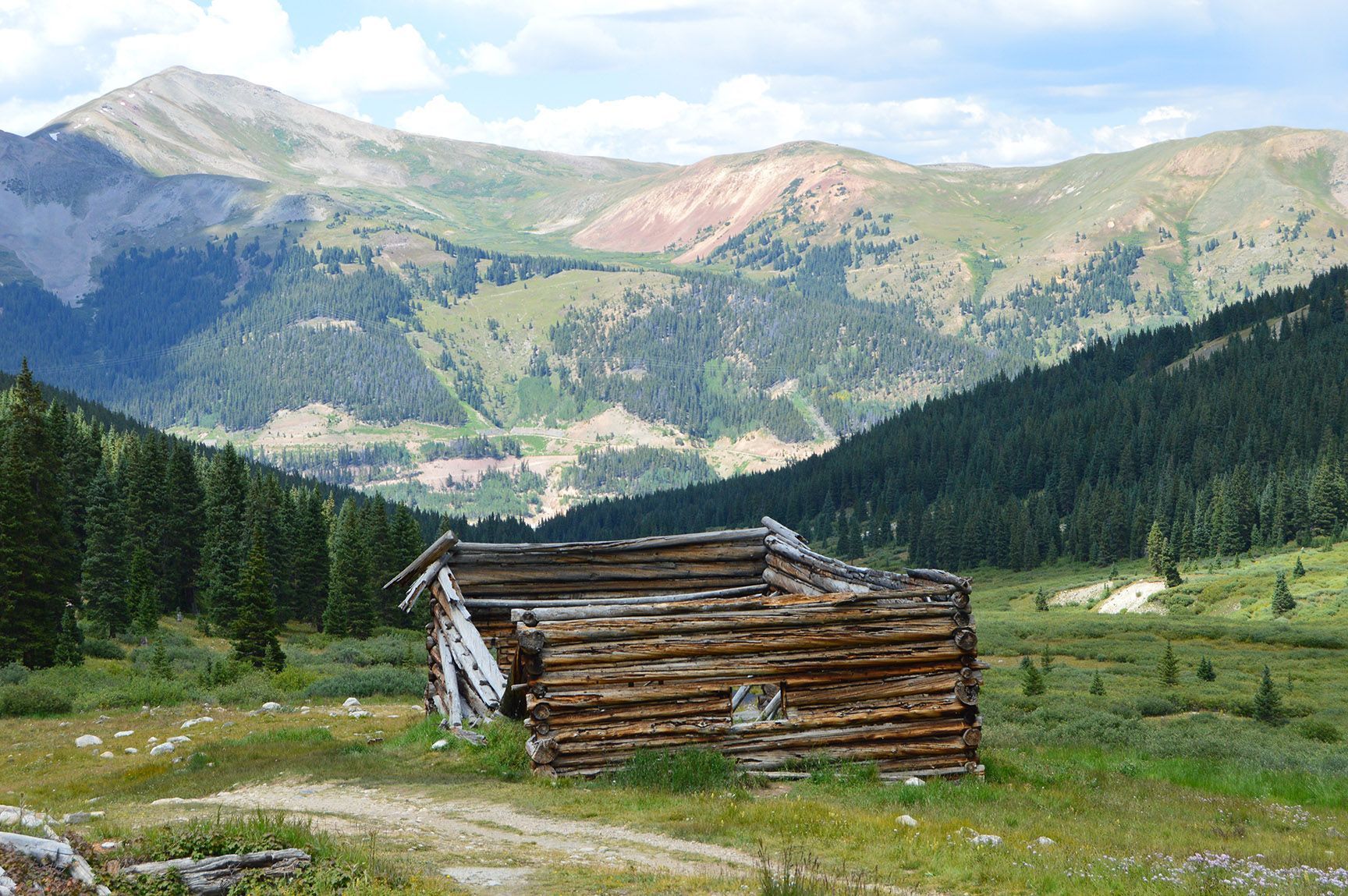 Small wooden cabin in a grassy mountain valley with pine forest and rugged peaks in the background, The ruins of an old cabin, Mayflower Gulch, Colorado