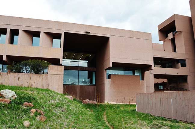 Modern tan building with large windows on a grassy hillside under cloudy sky