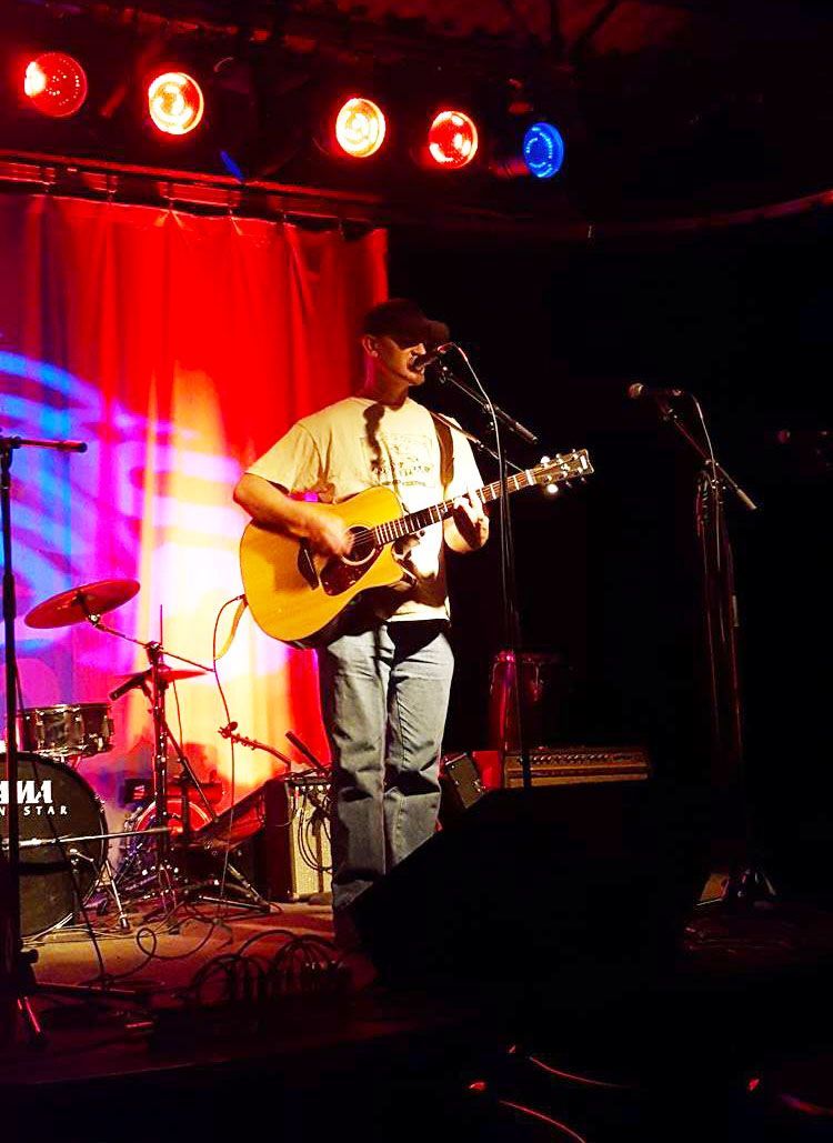 Musician playing acoustic guitar on a small stage with red lights and a drum set behind him