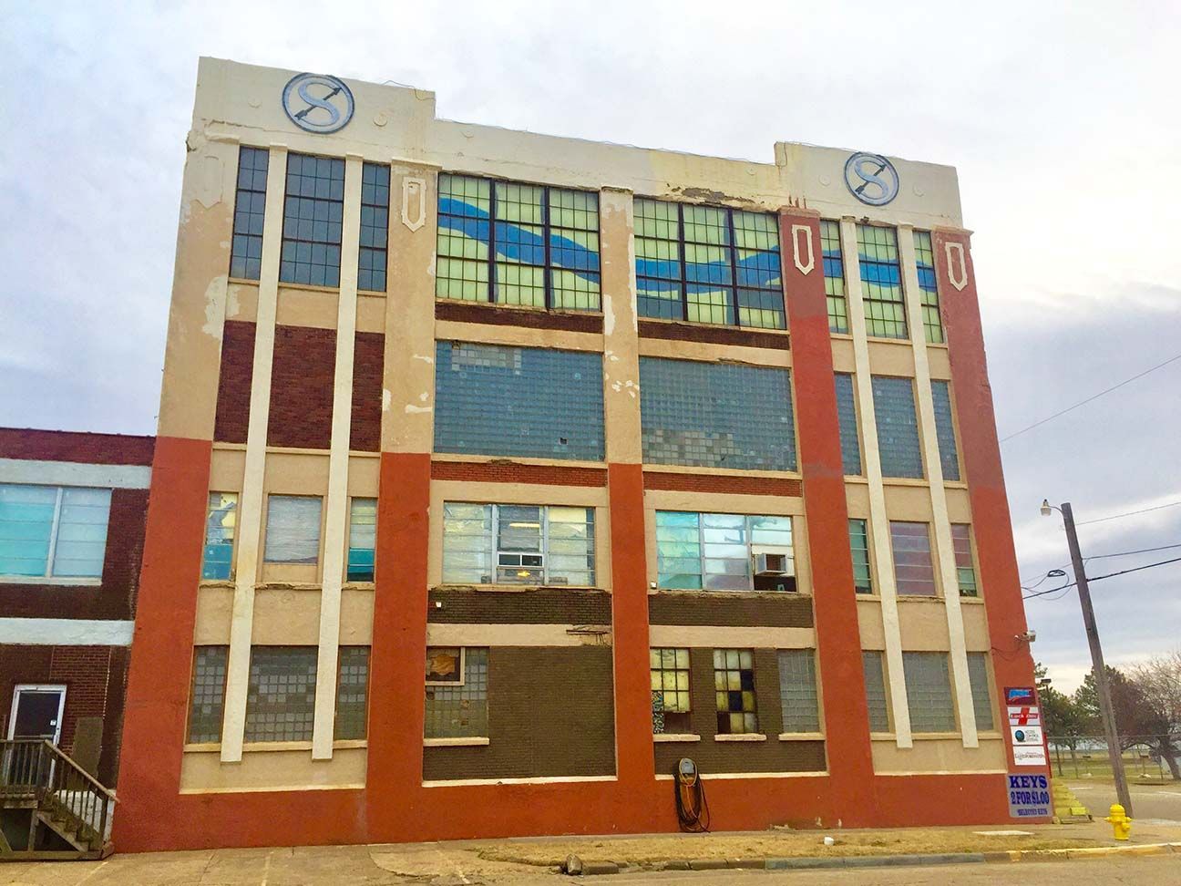 Multi-story brick building with large windows and clocks on the roofline under a cloudy sky