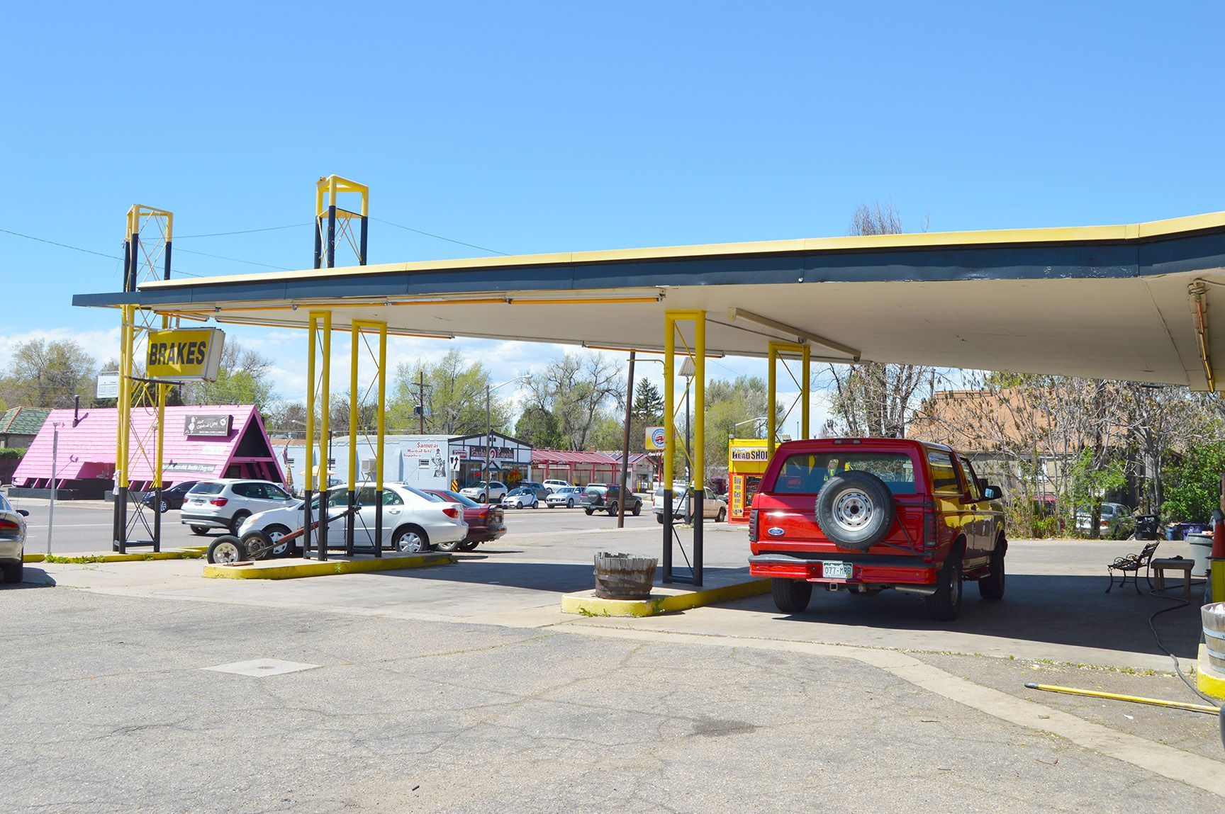 Red SUV under a yellow carport beside a parking lot with a pink van and other cars.
