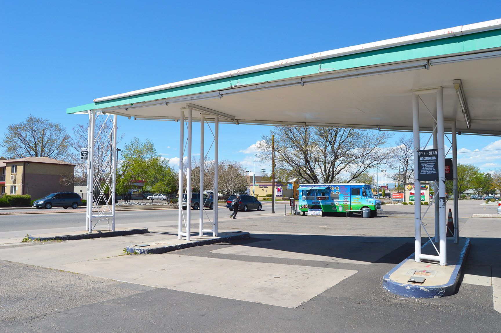 Abandoned gas station canopy with empty pumps and a few cars in the background under a clear blue sky
