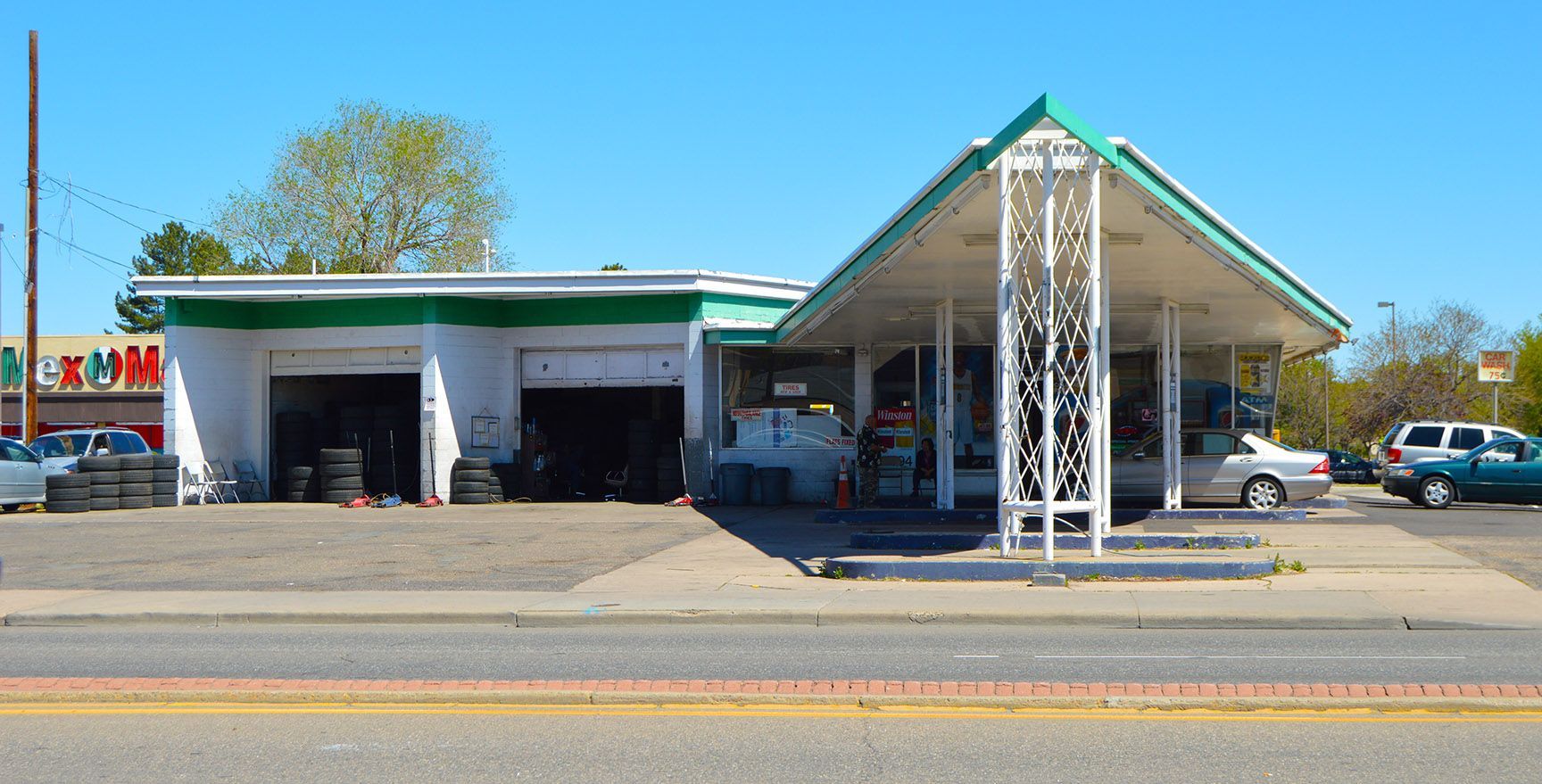 Small roadside building with green roof, open garage bays, and a covered entrance beside parked cars.