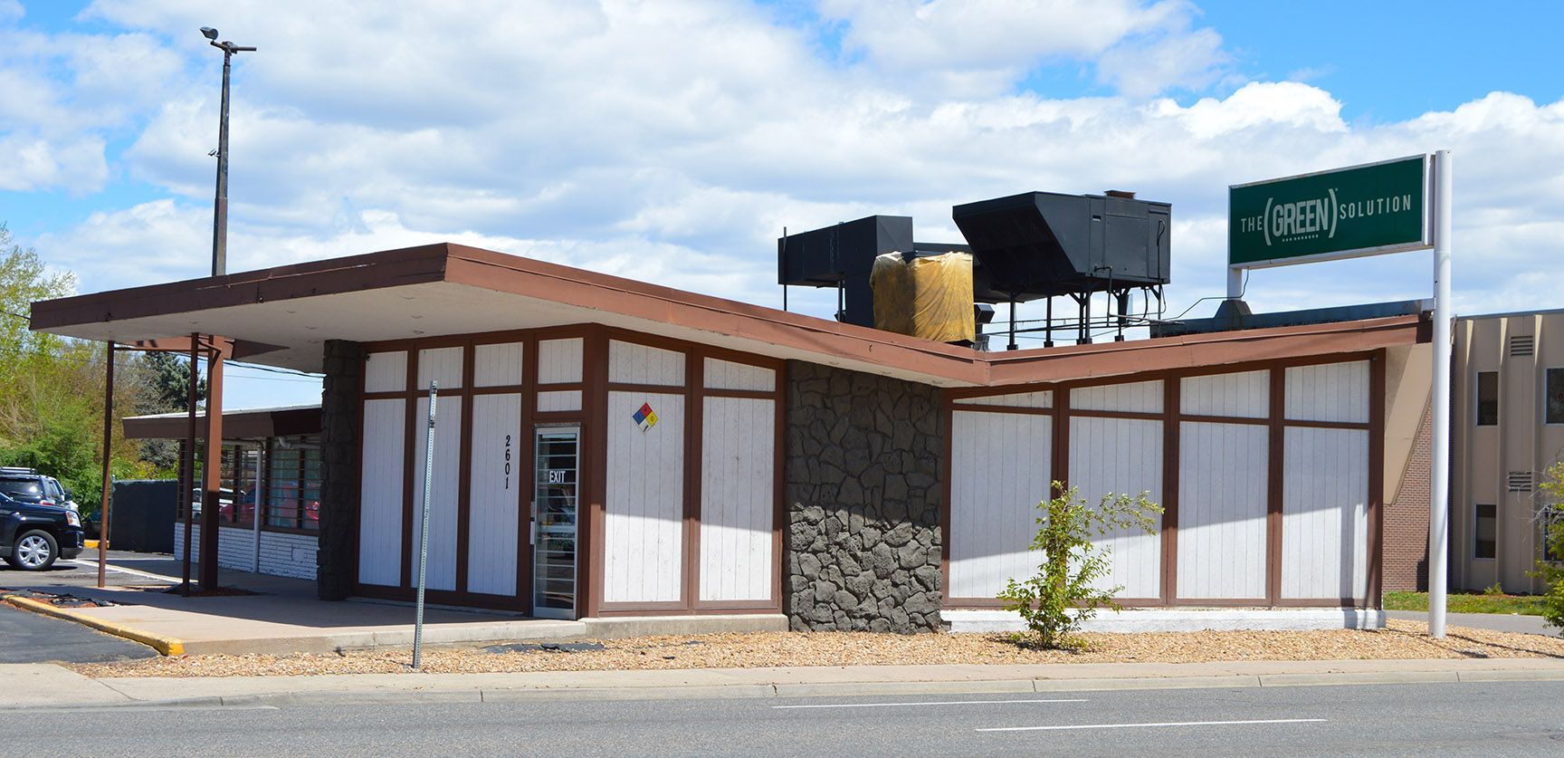 Small roadside building with a brown roof and white walls under a blue sky, near a green street sign