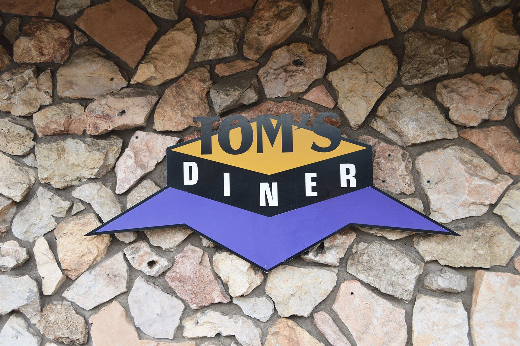 Tom’s Diner sign on a stone wall, with black and yellow lettering and a purple arrow-shaped banner.