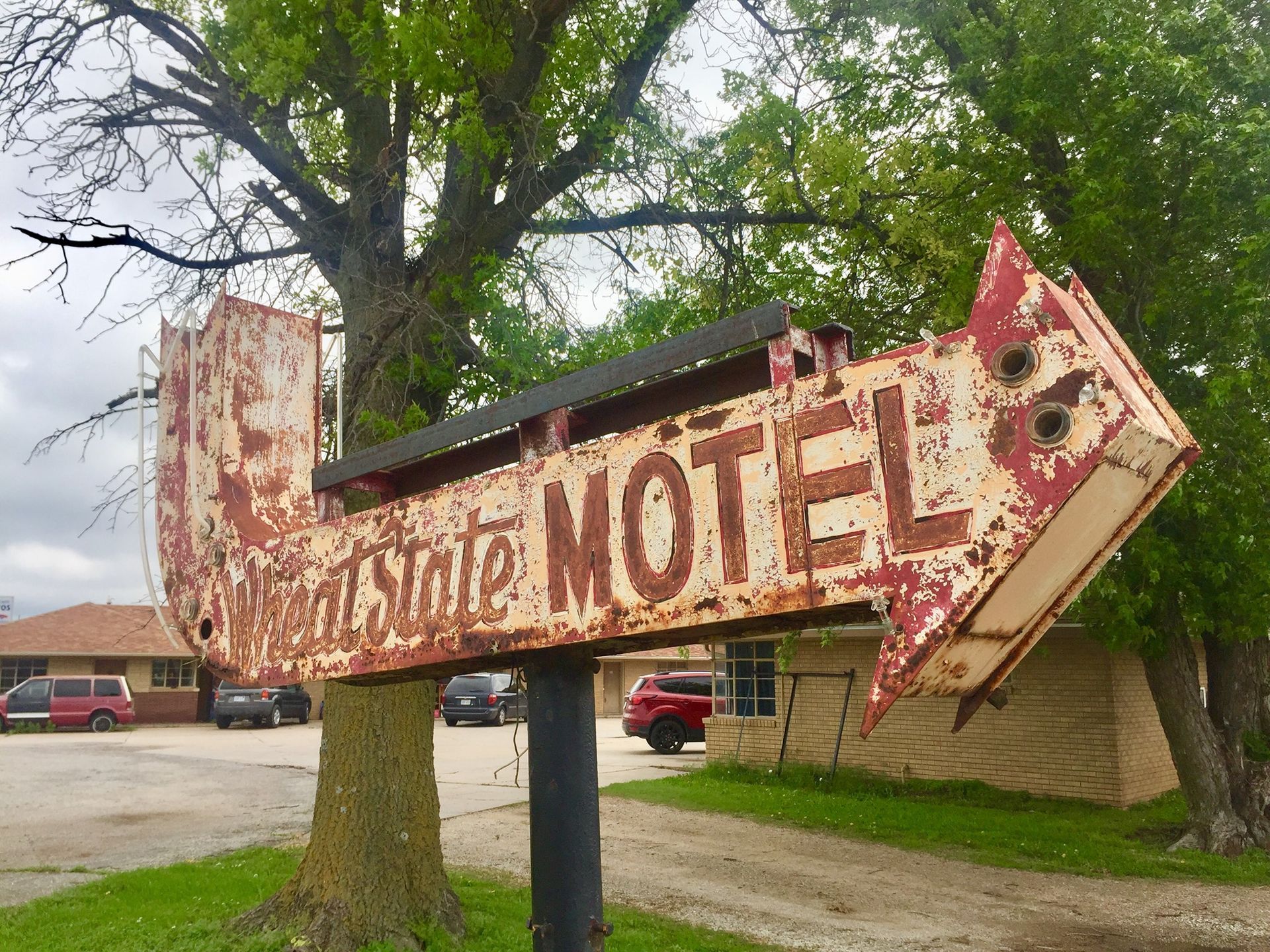 Rusty arrow motel sign with peeling paint in a roadside lot near trees and parked cars