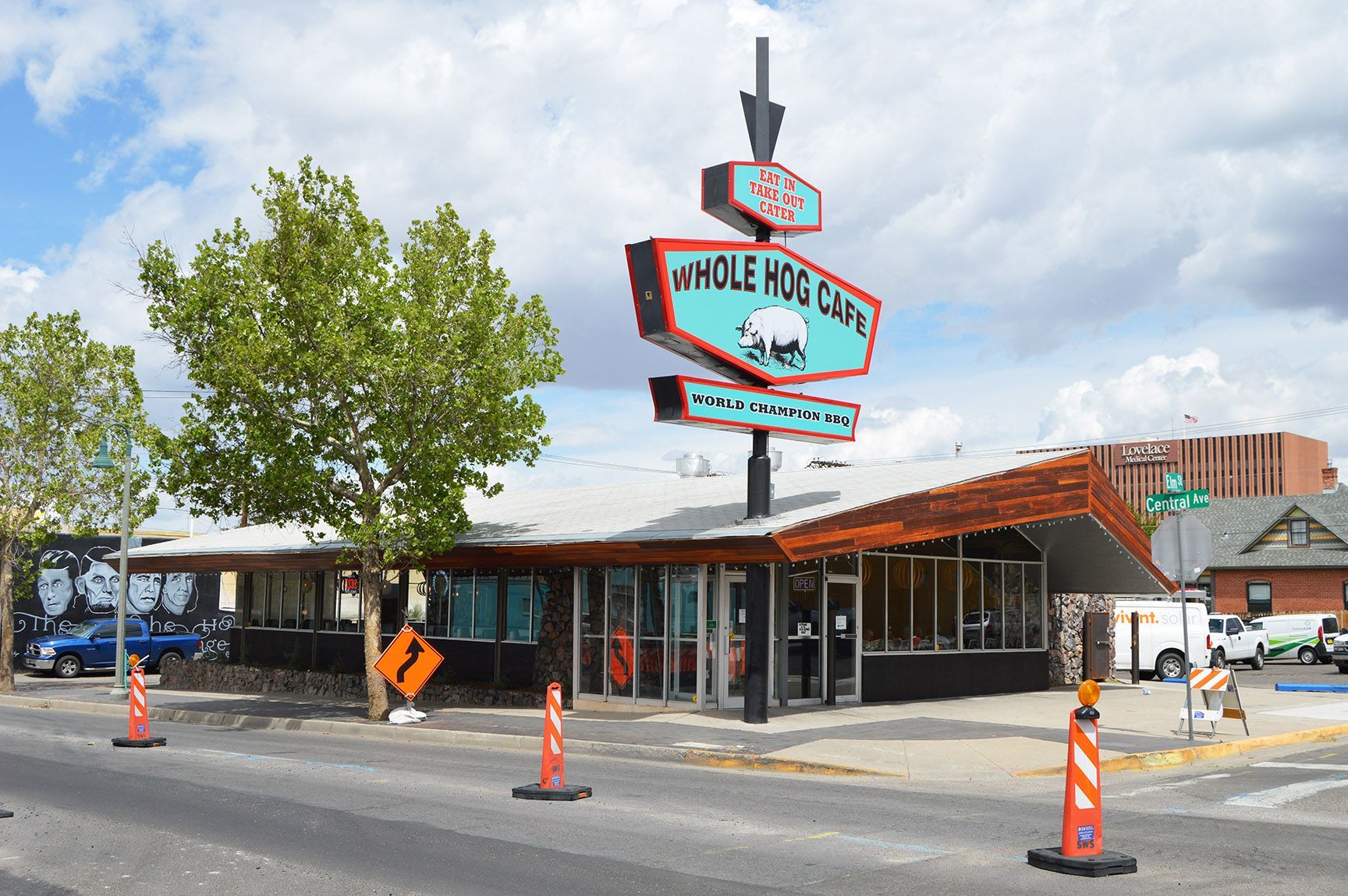 Street view of a diner with a retro neon sign, orange cones, and workers outside under a cloudy sky