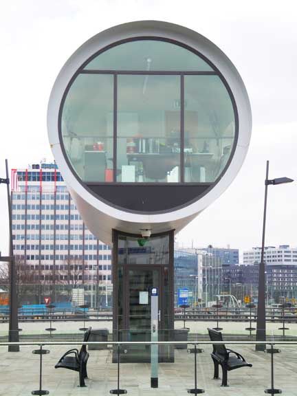 Round glass observation pod on a pedestal by the waterfront, with city buildings in the background
