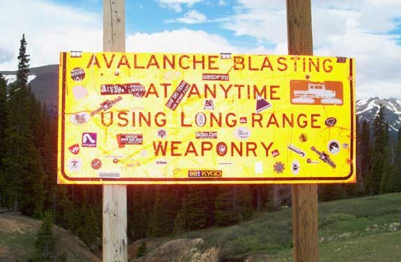 Yellow warning sign reading “AVALANCHE BLASTING AT ANYTIME USING LONG-RANGE WEAPONRY” beside a road