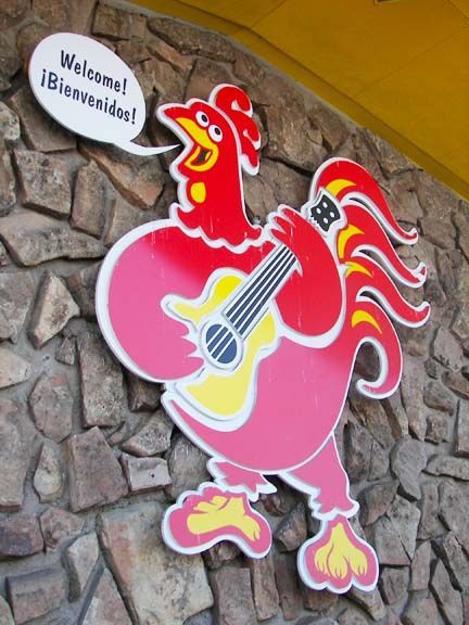 Pink cartoon rooster holding a guitar beside a “Willkomen!” welcome sign on a stone wall