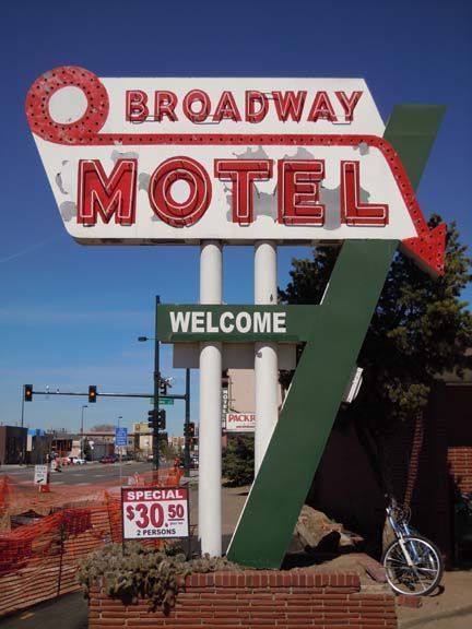 Broadway Motel neon sign with red letters and a green “WELCOME” arrow against a blue sky