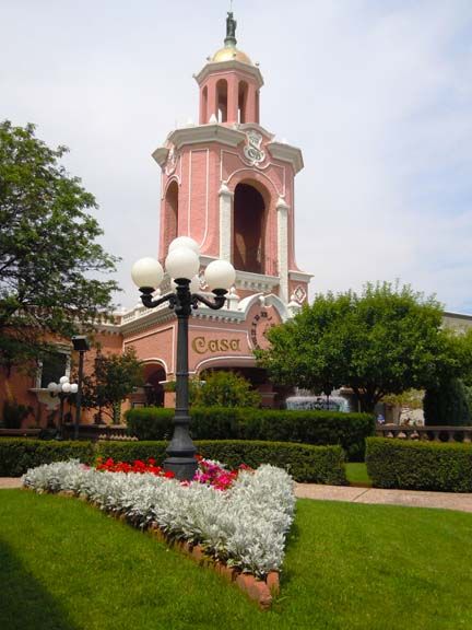 Pink tower with arches above a landscaped garden, white flowers, and a black lamppost under a cloudy sky