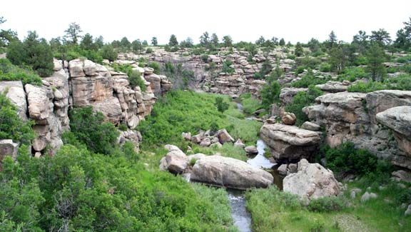 Rocky canyon with green shrubs, layered cliffs, and a small waterfall under an overcast sky