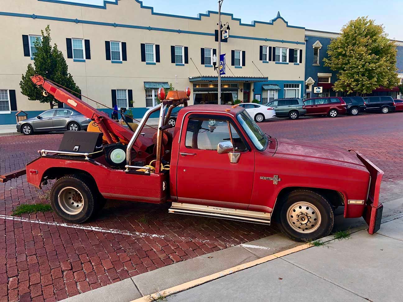 Red tow truck with raised boom parked on a brick street in front of buildings and parked cars, Lindsborg, Kansas