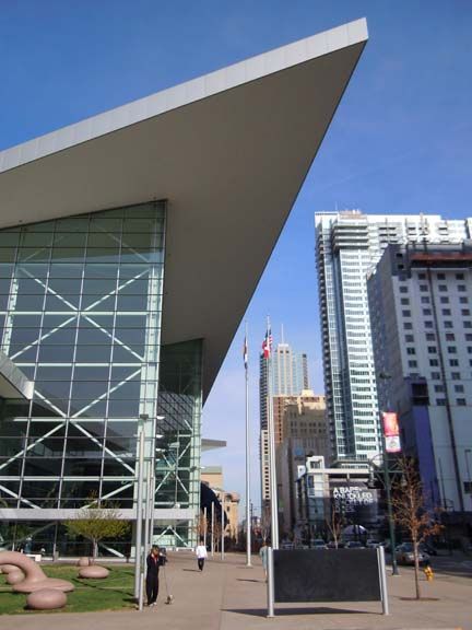 Modern glass building with a sharp overhang beside a city street and tall downtown towers