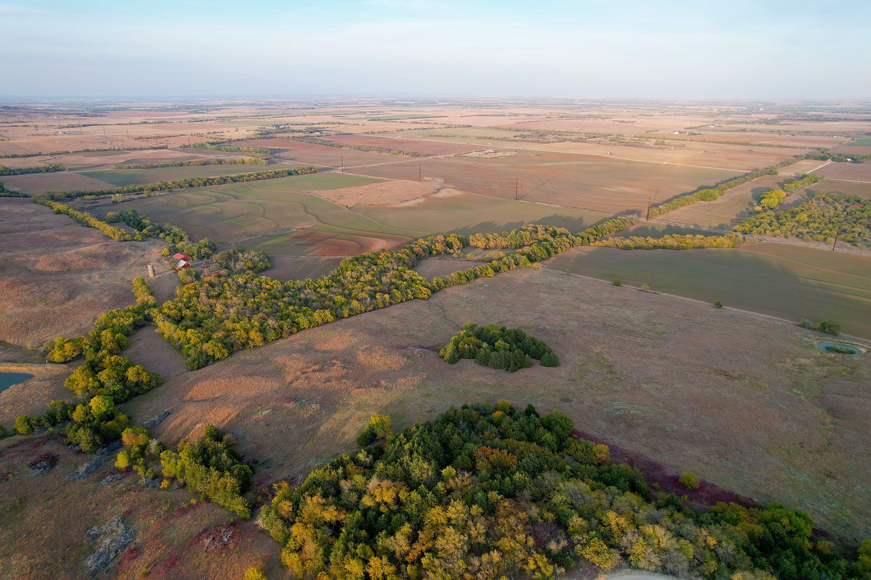 Aerial view of patchwork farmland with green tree lines and a winding river under a hazy sky