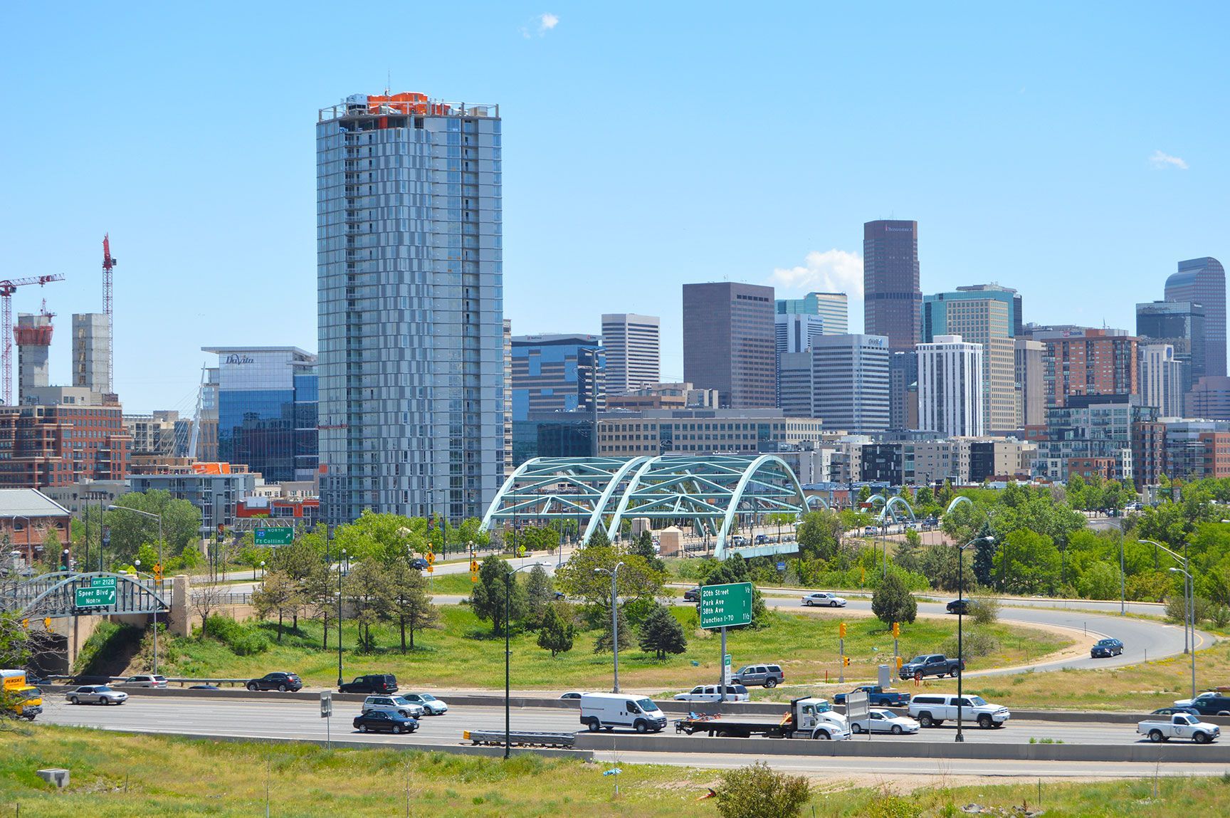 City skyline with a tall blue glass tower, mid-rise buildings, and a green park beside a road under a clear sky, Speer Blvd., Denver, Colorado, I-25, Interstate-25, cars driving, skyscrapers