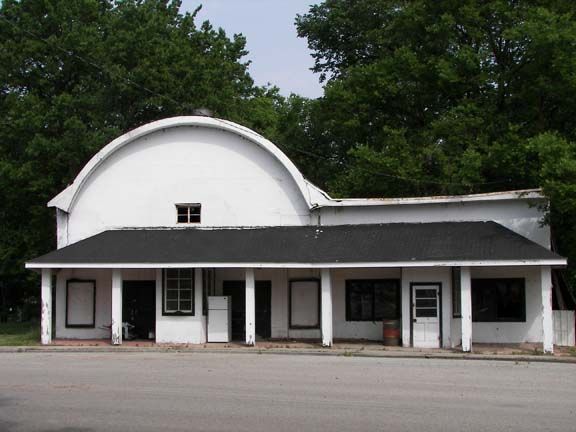 Old white building with a curved roof and front porch, surrounded by trees and an empty lot