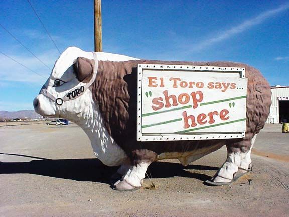 Large cow statue with a sign reading “El Toro says… shop here” by a roadside.