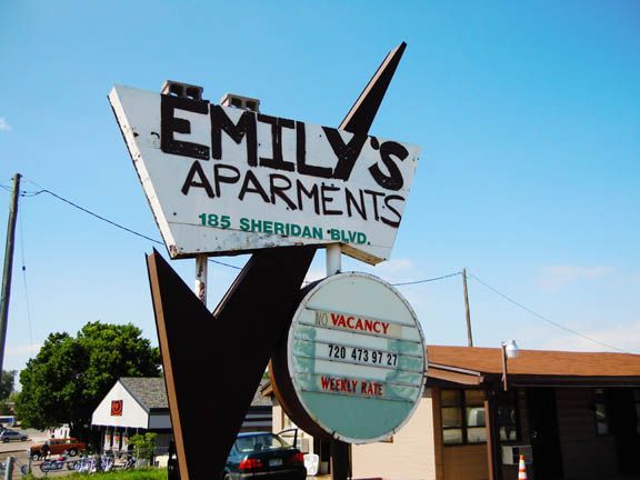 Motel sign reading “EMT Apartments” with “105 Sheridan Blvd” against a blue sky