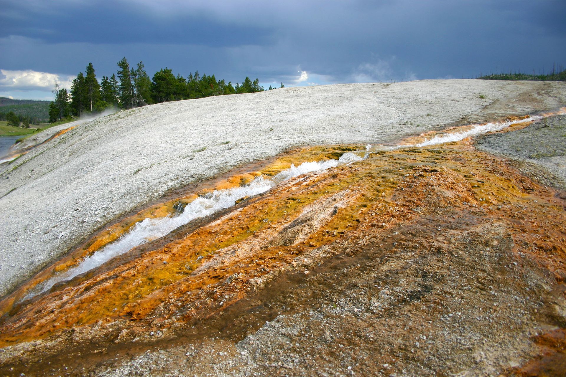 White and orange mineral-stained hillside under a cloudy sky with trees in the distance, Excelsior Geyser, Biscuit Basin, Yellowstone National Park, Wyoming