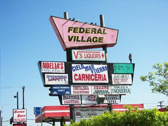 Roadside shopping plaza sign for Federal Village with multiple business names on stacked signs under a pink canopy.