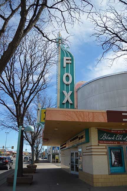 Fox Theater marquee with teal vertical sign on a city sidewalk, trees and storefronts nearby