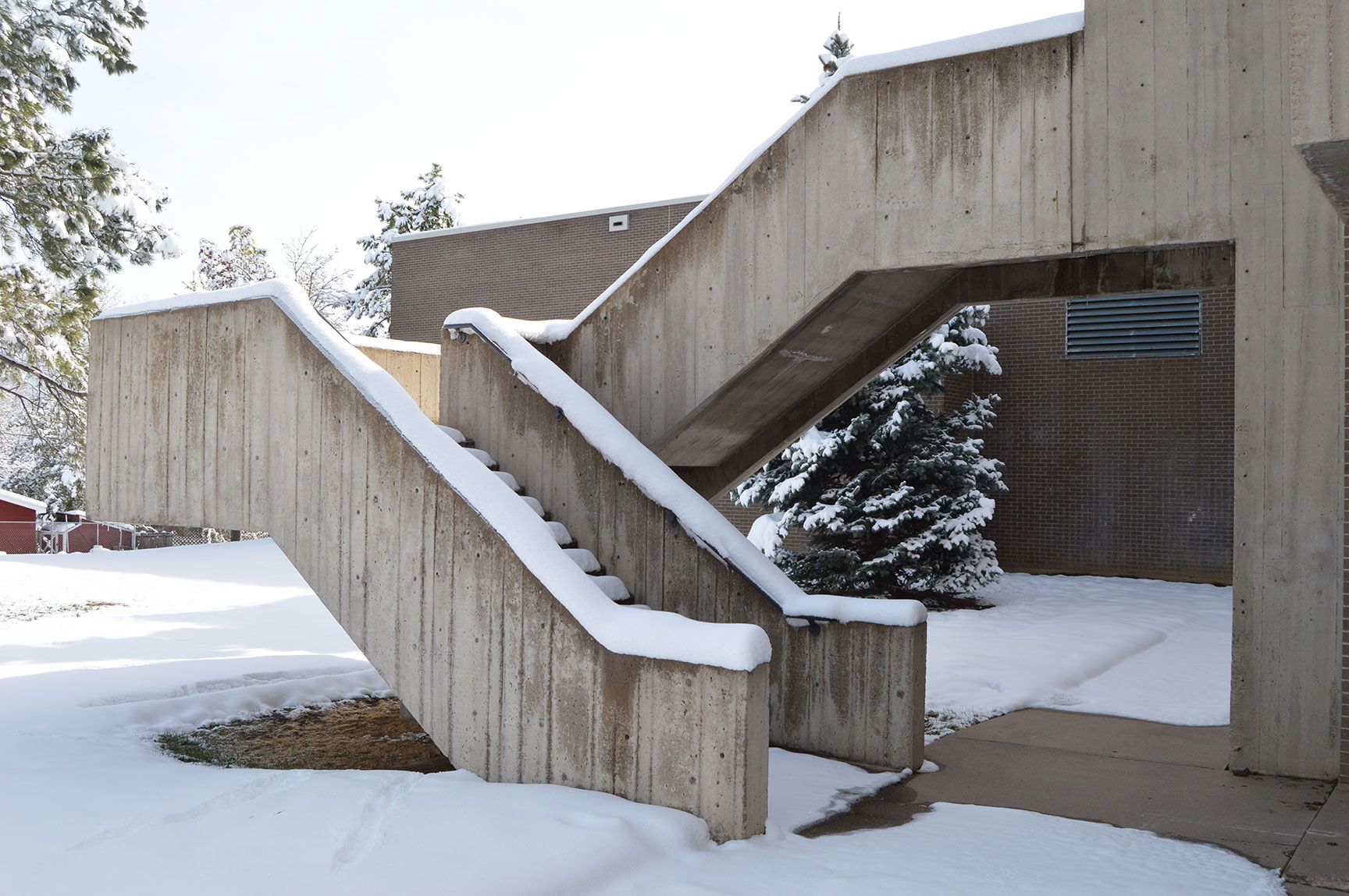 Snow-covered concrete stairway and ramp leading to a building entrance under an overhang.