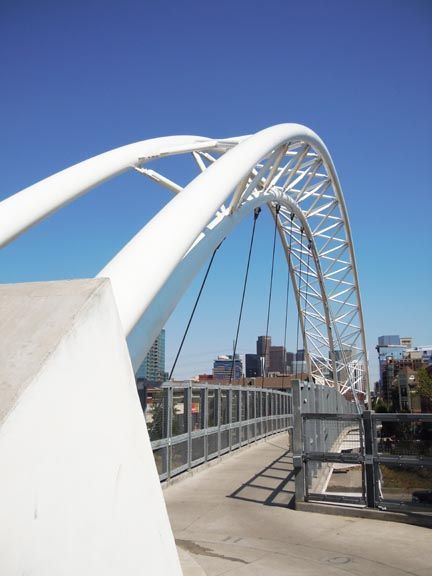 White arched pedestrian bridge with cables over a city walkway on a clear blue day