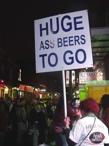 Person holding sign reading “HUGE ASS BEERS TO GO” at a nighttime street event