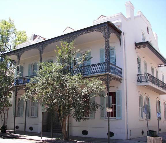Two-story cream house with blue wrought-iron balconies and trees in front