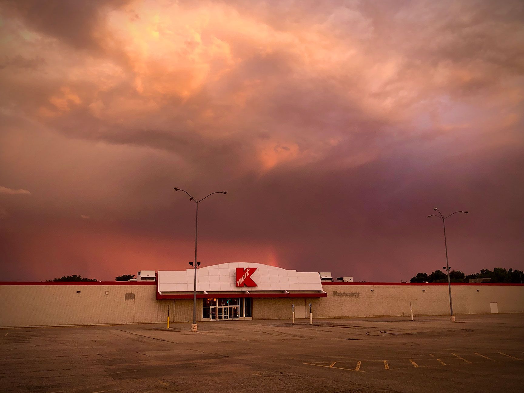 Kmart store beneath a dramatic orange storm sky at dusk, with empty parking lot and distant birds.