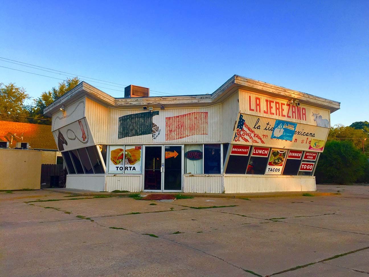 Small roadside storefront with a faded white and red facade and Spanish signs, under a blue sky.