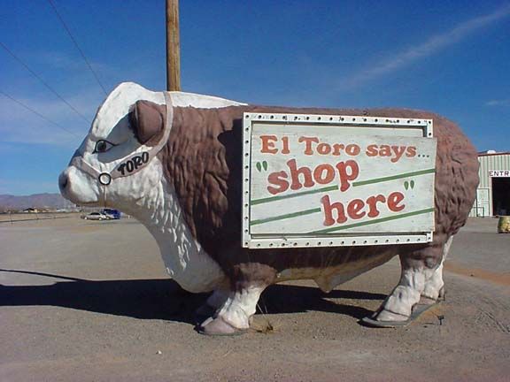Bull statue with a sign reading “El Toro says... shop here” in a parking lot under blue sky