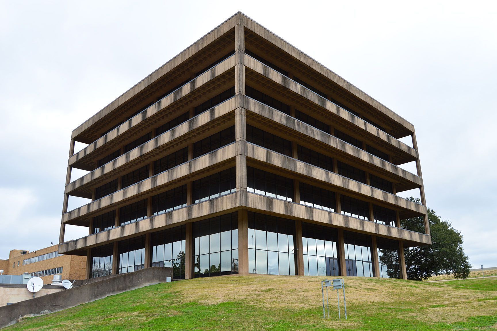Four-story brown office building with glass windows on a grassy slope under a cloudy sky