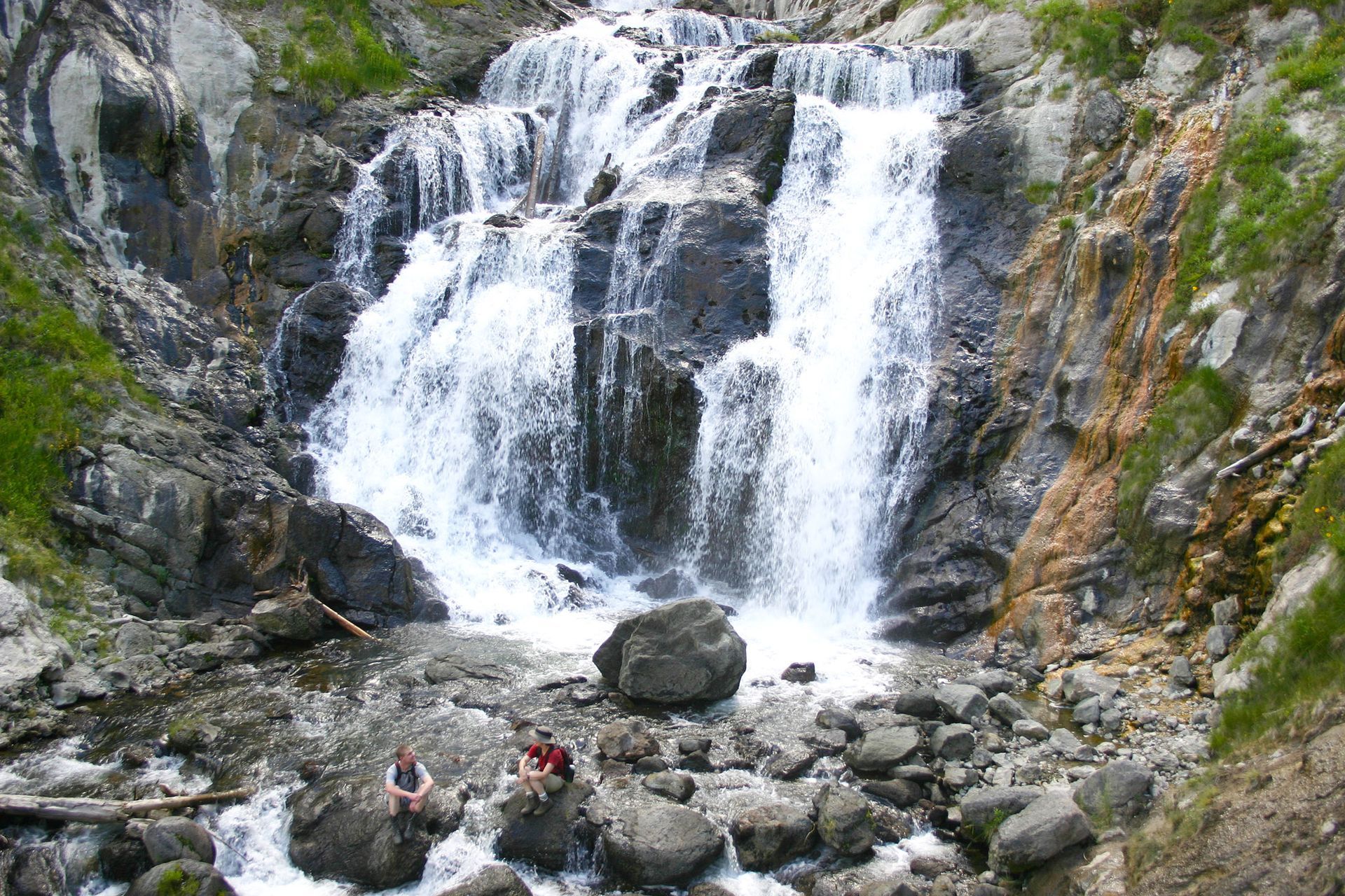 Waterfall cascading over rocky cliff into a boulder-strewn pool with a few hikers below, Mystic Falls, Yellowstone National Park, Wyoming