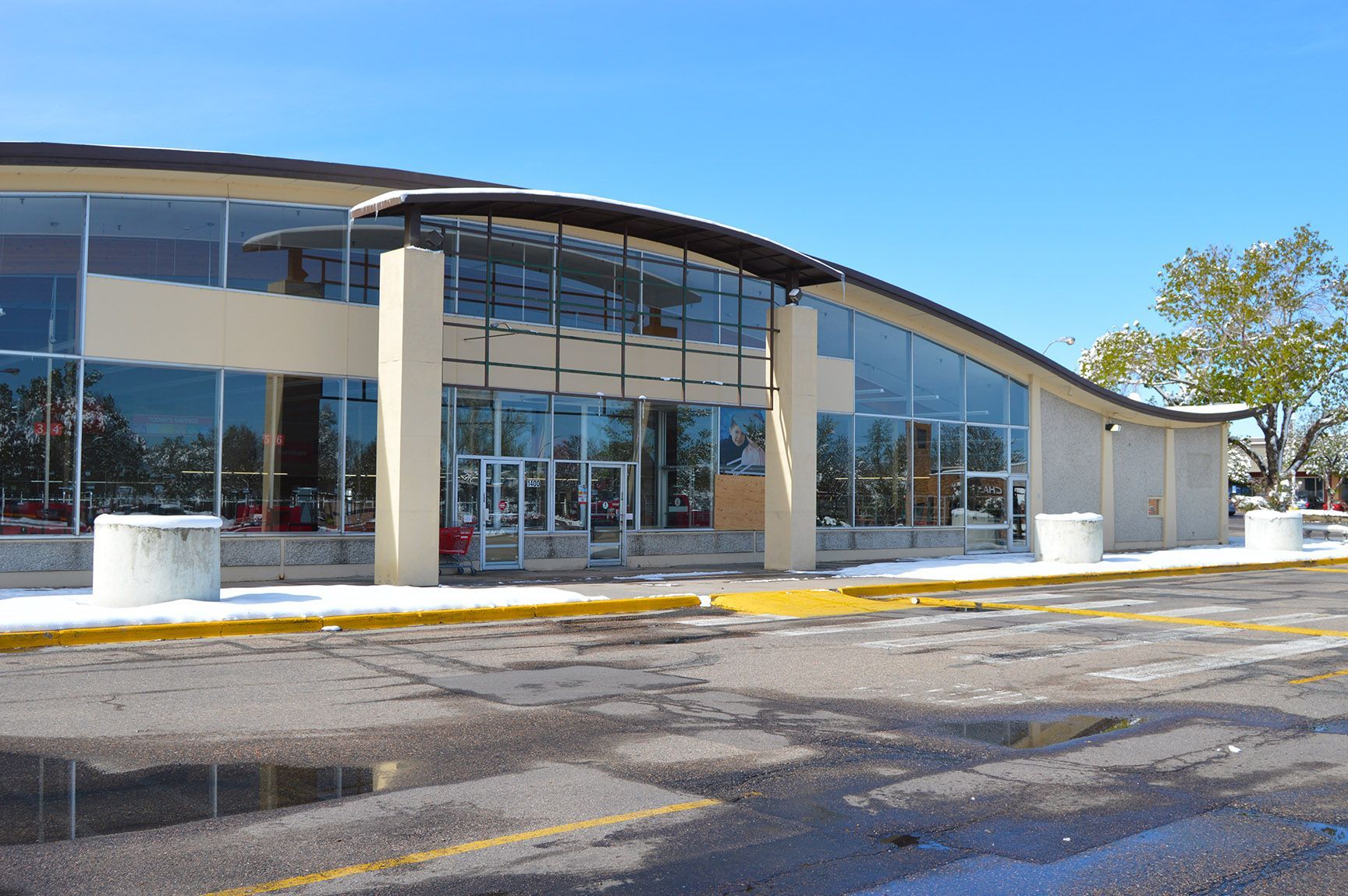 Modern glass-fronted building entrance with curved roof, columns, and empty parking lot under a clear blue sky