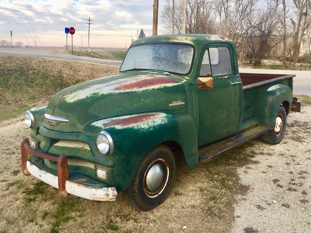 Old green vintage pickup truck parked on a gravel roadside beside bare trees at dusk, Chevrolet pickup truck, Salina, Kansas