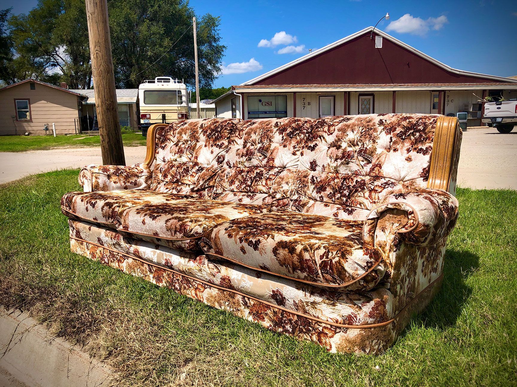 Moldy couch on a lawn in front of a house, with a driveway and trees in the background
