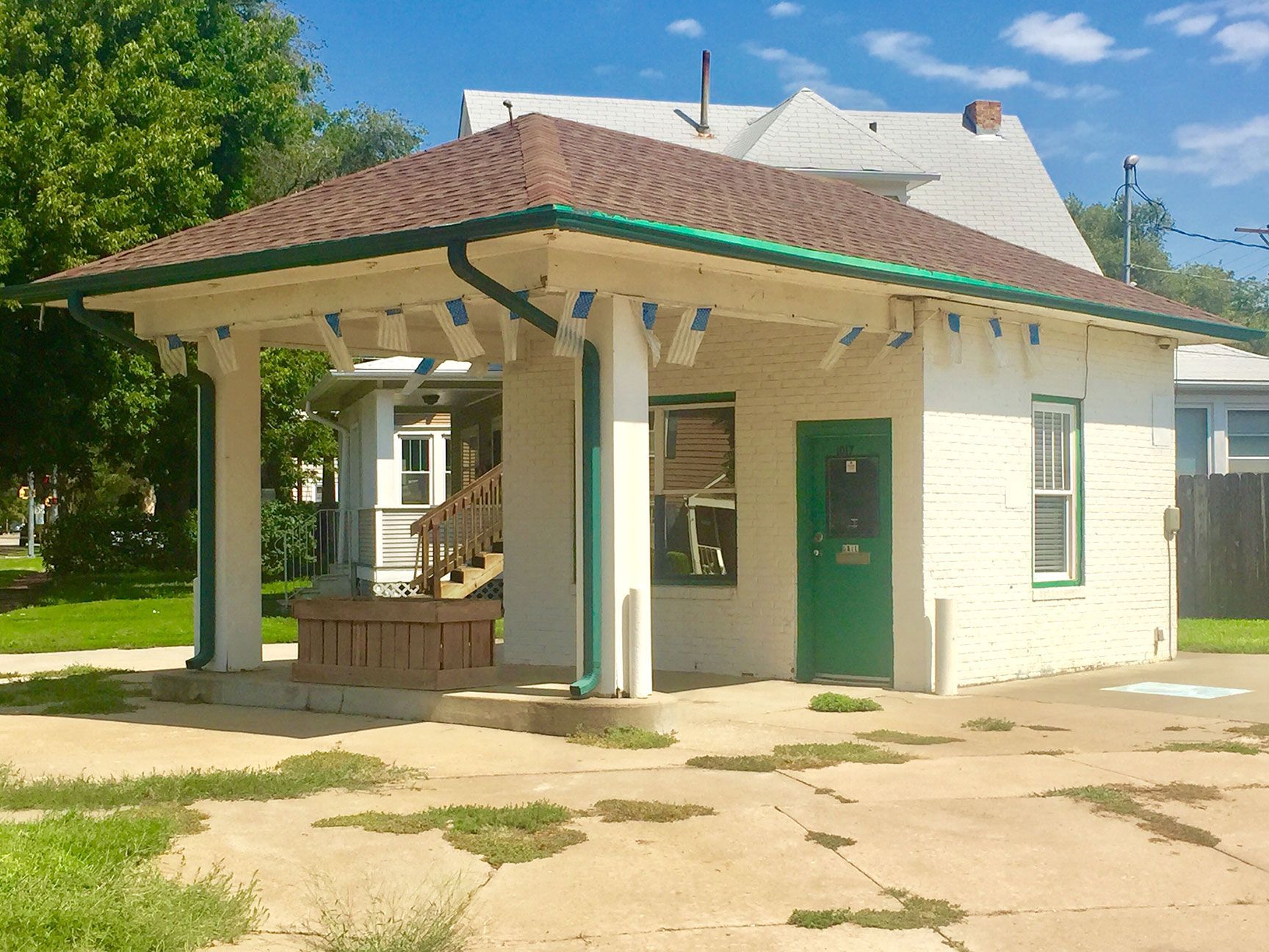 Small white building with a green door and brown roof, seen on a sunny day.