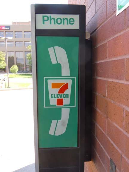 7-Eleven pay phone booth with green signage beside a brick wall